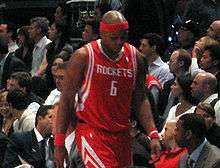 A black man wearing a red and white basketball uniform and matching red headband walks to the bench during a basketball game with his head down.