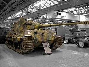 A large, turreted tank with dull yellow, green and brown wavy camouflage, on display inside a museum. The tracks are wide, and the frontal armour is sloped. The long gun overhangs the bow by several meters.