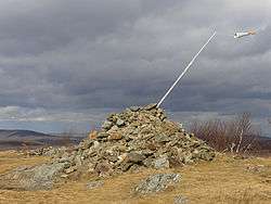 A pile of stones in a grassy clearing in late autumn. A pole with a small windsock protrudes from its right