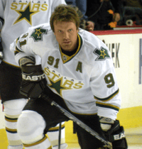 Hockey player in white Dallas Stars uniform, his helmet off. He leans forward and low, as if he were preparing to speed skate across the ice.