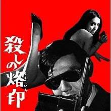 Joe Shishido holds a gun against his face. Annu Mari lays on her stomach and looks back over her shoulder aiming a gun behind him.