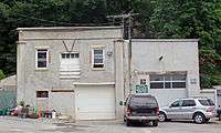 A gray building with two sections, both with large garage doors. A small green sign on the front of the smaller section, to the right, indicates that it is a registered New York State motor vehicle repair shop. Two cars are parked in front of that section, and behind the building is woods