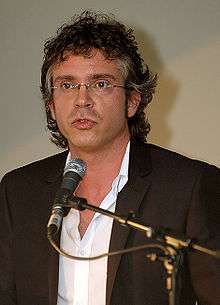 Man in white shirt and dark jacket speaking at a microphone