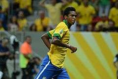 Man with dark skin and short black hair, wearing a football kit composed of a yellow shirt with green collar and blue shorts, running in front a filled stadium stand.