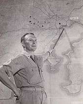 Black and white photo of a middle-aged man wearing military uniform pointing a stick at a map of the Tokyo region of Japan