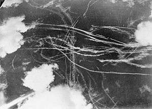 A picture of a piece of sky with several clouds and many condensation trails caused by many aircraft. Each trail curves around the other indicated an air battle