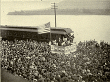 A dramatic political scene. Beside a river stands a podium, on which a flagpole flies a huge American flag. Beneath the flag stands a candidate in a dark suit addressing an impressive crowd that takes up most of the photograph. Not only the quayside, but a ferry beside it on the water are packed full of people listening intently.