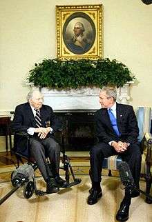 Frank Buckles in a wheel chair is talking to George W. Bush. In the background, above their heads are a plant decoration and a portrait of George Washington.