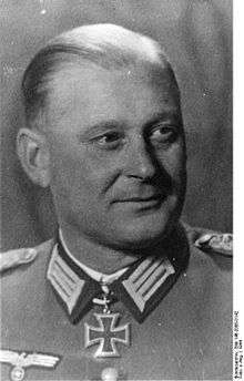 The head of a man, shown in semi-profile. He wears a military uniform with a military decoration in shape of an iron cross displayed at the front of his shirt collar. His hair is combed to back.