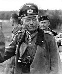 Guderian is seen in the field. He wears a military uniform and an Iron Cross displayed at the front. He is wearing a greatcoat and has binoculars around his neck.