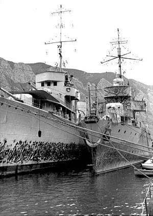 two naval ships side by side alongside a dock with mountains in the background