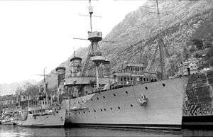 one larger naval ship moored alongside two small ships with mountains in the background