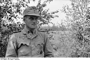 A black-and-white photograph of a man wearing a military uniform, field cap and a neck order in shape of an Iron Cross standing in front of a tree.