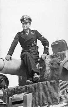 A man, wearing dress uniform and a cap, sits on top of a tank barrel