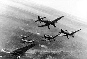 a black and white photograph of seven aircraft in flight over a smoky battleground