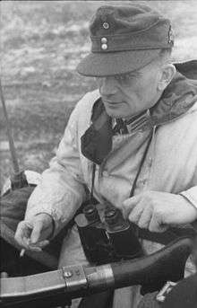 Black-and-white portrait of a man in semi profile wearing a field cap, coat with binoculars suspended from his neck. He is holding a cigarette in his right hand.