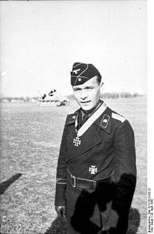 Black-and-white portrait of a standing man wearing a cap and black military uniform with an Iron Cross displayed at his neck.