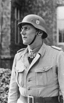 A man in semi profile wearing a military uniform, steel helmet and a neck order in the shape of a cross.