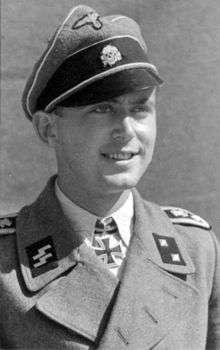A man wearing a military uniform, peaked cap and a neck order in the shape of a cross. His cap has an emblem in shape of a human skull and crossed bones.