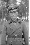 A man wearing a military uniform and coat, peaked cap and a neck order in the shape of a cross. His cap has an emblem in shape of a human skull and crossed bones.