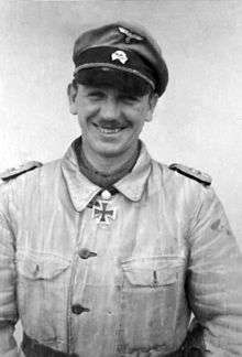 A smiling man wearing a military uniform, peaked cap and neck order, in the shape of a cross.