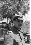 A man in semi profile standing in front of a tree and wearing a military uniform, peaked cap and a neck order in the shape of a cross. His cap has an emblem in shape of a human skull and crossed bones.