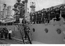 Men in military uniforms walk down stairs on a large warship as dozens of sailors look on and salute.