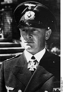 The head of a young man, shown in semi-profile. He wears a peaked cap, naval military uniform with a military decoration in shape of an iron cross displayed at the front of his shirt collar.