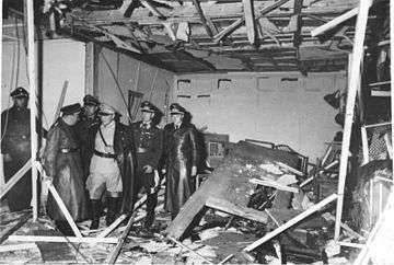 A black-and-white photograph of six men in military uniforms while surveying a shattered conference building.