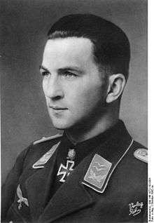 The head of a young man, shown in semi-profile. He wears a military uniform with an eagle above his right breast pocket, an iron cross is displayed at the front of his shirt collar. His hair is dark, short and combed to the back, his nose is long and straight, and his facial expression is determined; looking to the right of the camera.