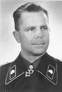 The head a man, shown from the front looking slightly to his left. He wears a black military uniform, a dark shirt with an Iron Cross displayed at the front of his shirt collar. On the collar of his uniform are two skull insignias. His hair appears dark and is combed back, his nose is scared, his facial expression is a determined.