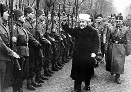 a man wearing Muslim mufti clothing holding his hand up in salute as he and a group of SS officers inspect a line of soldiers