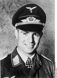 A smiling man wearing a military uniform, peaked cap, and an Iron Cross displayed at the front of his uniform collar.
