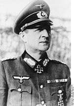A man wearing a military uniform, peaked cap and neck order in the shape of a cross.