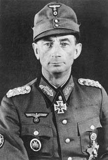 The head and shoulders of an elderly man, shown from the front. He wears a field cap and a military uniform displaying various military decorations on breast pockets, right upper sleeve, and an Iron Cross displayed at the front of his shirt collar. His facial expression is a determined and confident smile; his eyes look into the camera.
