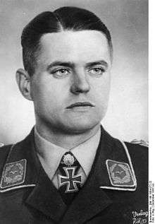 The head and shoulders of a young man, shown from the front. He wears a military uniform, with an Iron Cross displayed at the front of his white shirt collar. His facial expression is a determined and a grim smile; his eyes are looking to the left of the camera.