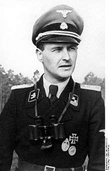 A man wearing a military uniform, peaked cap and a pair of binoculars around his neck. His cap has an emblem in shape of a human skull and crossed bones.