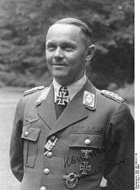 A man wearing a military uniform and neck order, in the shape of a cross. He has short hair that is combed back and a determined facial expression.