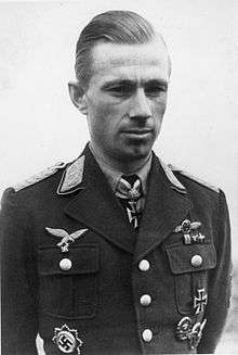 Black-and-white photograph showing the face and upper body of a young man in uniform, his hands behind his back. His hair appears blond and combed to the back. The front right of his jacket bear eagle-and-swastika emblems; the front left of his jacket and the front of his shirt collar bear Iron Cross decorations, black with light outline. He is looking at the camera, his facial expression is secluded.