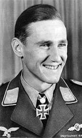 The head of a young man, shown in semi-profile. He wears a military uniform with a military decoration in shape of an iron cross displayed at the front of his shirt collar. His hair is dark and short and combed to back, his nose is long and straight, he is smiling broadly and looking to the left of the camera.