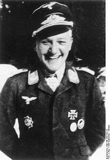 A smiling man wearing a peaked cap, military uniform with an Iron Cross displayed at the front of his uniform collar.