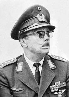The head and shoulders of a man, shown in semi-profile. He wears a peaked cap and a military uniform with military decorations. His face is scared and his eyes are hidden behind glasses.
