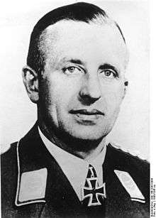 The head of a man, shown in semi-profile. He wears a military uniform with a military decoration in shape of an iron cross displayed at the front of his shirt collar. His hair is dark and combed to back.