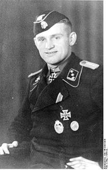 A man wearing a military uniform, side cap and neck order in the shape of a cross. His cap has an emblem in shape of a human skull and crossed bones.