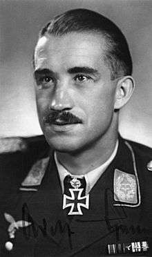 The head and shoulders of a young man, shown in semi-profile. He wears a military uniform with various military above his left breast pocket and an Iron Cross displayed at the front of his shirt collar. On his upper lip is a moustache, his hair is dark and short and combed back, his facial expression is a determined and confident smile; his eyes gaze into the distance.