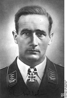 The head a man, shown from the front. He wears a military uniform, a white shirt with an Iron Cross displayed at the front of his shirt collar. His hair appears dark and is combed back, his facial expression is a determined; his eyes are looking into the camera.