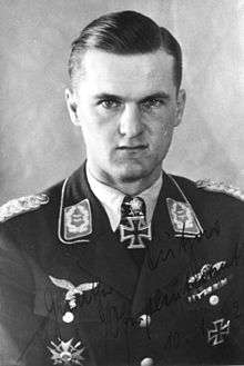 The head and shoulders of a young man, shown in semi-profile. He wears a military uniform with an Iron Cross displayed at the front of his shirt collar. His hair is short and combed to his right, his nose is short, and his facial expression is determined; looking into the camera.