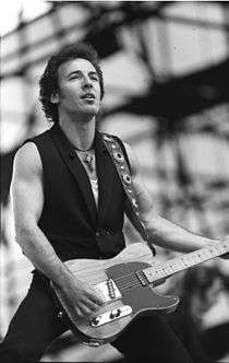 Black and white image of a man holding a guitar, wearing a dark vest and a cross hanging from a necklace