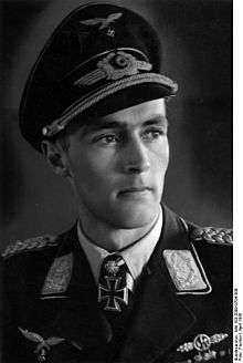 The head and shoulders of a young man, shown in semi-profile. He wears a peaked cap and a military uniform with an Eagle above his right and a military decorations above left breast pocket, and an Iron Cross displayed at the front of his shirt collar. His facial expression is a determined; his eyes are looking into the distance to the left of the camera.