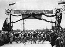 A black-and-white photograph showing a group of cyclists, with a banner above them